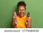 Unhealhty food and weight loss concept. Positive smiling woman keeps eyes closed and laughs, holds strawberry popsicle and chocolate ice cream, isolated on green background. Summer time, eating