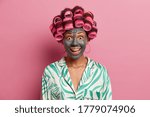 Happy joyful woman visits hairdressing and spa salon, makes perfect hairstyle and applies clay face mask, wears pyjama, has surpised expression, isolated on pink background. Female going on date