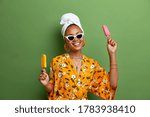 Carefree positive dark skinned woman holds delicious ice cream, popsicles on stick, has fun during summer time, wears stylish sunglasses, yellow robe, wrapped towel on head, has sweet tooth.