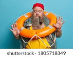 Small photo of Scared seaman raises palm, stares bugged eyes, afraids of smimming, poses with orange inflated lifebuoy, fishing net, being seasick, isolated on blue background. Fearful member of deck department