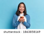 Beautiful Asian woman holds modern cellular glad to surf in social networks smiles positively dressed in casual jumper poses against blue background. Technology and online communication concept