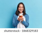 Smiling charming brunette Asian woman uses mobile phone happy texting in social networks addicted to modern technologies wears casual jumper isolated over blue background. Online communication