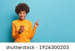 Small photo of Horizontal shot of happy dark skinned Afro American woman enjoys mobile communication and modern technologies poses against blue background points away on free space for your advertising content