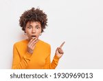 Horizontal shocked speechless woman with Afro hair keeps jaw dropped indicates away on blank space says check out something unusual wears long sleeved orange jumper isolated over white background