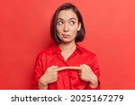 Small photo of Hesitant indecisive pretty Asian woman keeps index finger opposite feels doubtful wears shirt focused above poses against red background thinks about something. Let me think for few minutes.