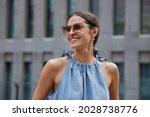 Pleased European woman with dark hair smiles broadly wears sunglasses blue dress enjoys summer walk in urban setting glad to meet friend outside expresses positive emotions returns glad after shopping