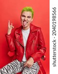 Small photo of Sassy punk girl sticks out tongue shows heavy metal sign gesture feels cool has trendy green hairstyle wears leather jacket sits on chair isolated over red background enjoys rock music. Youth concept