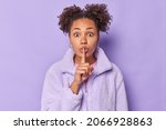 Small photo of Cute shocked young woman makes silence gesture presses index finger over lips shows hush sign says shh dressed in outerwear spreads gossips isolated over purple background. Body language concept