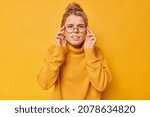Small photo of Young pretty woman has bad eyesight tries to see something far away focused into distance tries new spectacles for good vision wears jumper isolated over vivid yellow background. Scrupulous gaze