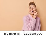 Small photo of Happy carefree woman with hair gathered in bun touches face gently looks away smiles broadly wears knitted sweater isolated over beige background blank copy space for your promotional content