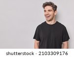 Small photo of Horizontal shot of handsome cheerful man looks happily away smiles broadly wears casual black t shirt isolated over white background with blank copy space being in good mood laughs at something