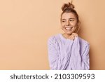 Small photo of Horizontal shot of happy European woman looks dreamy away keeps hands together smiles broadly has good mood wears knitted sweater poses against beige bacground with blank copy space for promo