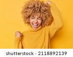 Small photo of Overjoyed curly haired woman makes yes gesture feels excited and triumphant exclaims from joy wears casual sweater poses over yellow studio background celebrates success. Yes I achieved my goal