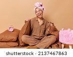 Glad young woman sits in lotus pose on soft bed wears pajama wrapped towel on head laughs happily away feels relaxed after sleeping covered with blanket surrounded by pillows and alarm clock