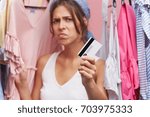 Small photo of Sad upset girl shoppaholic standing at rack of stylish clothes in store, holding credit card, feeling worried and unhappy after she overspent money on buying too much items. Consumerism and addiction