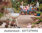 Wooden patio in the garden with a grill standing in the front and happy young people gathered around a table full of food during summer brake meeting