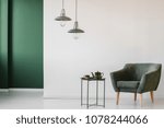 Upholstered, dark armchair and an industrial side table with a tea kettle and cup in a minimalist living room interior with a green corner