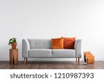 Comfortable couch with orange and red pillow in spacious living room interior, real photo with copy space on the empty white wall