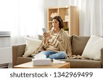 technology, health and cold concept - sad sick woman in blanket using smartphone at home