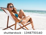 Happy young woman relaxing on deck chair at beach while looking at camera. Mature woman with red hair wearing sunglasses and blue bikini enjoying vacation at beach. Sunbathing and relaxing at sea.