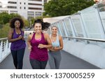Happy young curvy women jogging together on city bridge. Healthy girls friends running on the city street to lose weight. Group of multiethnic oversize women running with building in the background.
