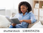 leisure, literature and people concept - smiling african american woman reading book at home