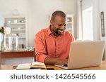 Smiling black man using laptop at home in living room. Happy mature businessman send email and working at home. African american freelancer typing on computer with paperworks and documents on table.
