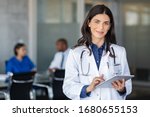 Portrait of beautiful mature woman doctor holding digital tablet and looking at camera. Confident female doctor using digital tablet with colleague talking in background at hospital. Latin nurse.