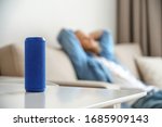 Small portable wireless speaker assistant on table with man lounge on sofa listening to music. Young relaxed guy enjoys mini bluetooth stereo gadget for sound digital assistance at smart home concept.