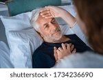 Woman checking fever temperature of senior man lying on bed. Mature husband resting at home feeling symptoms of the flu while wife checking fever by touching forehead. Close up face of old sick man.