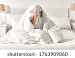 morning, comfort and people concept - young woman with cup of coffee sitting in bed under blanket at home bedroom