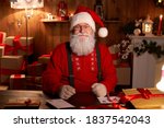 Portrait of happy old bearded Santa Claus wearing hat, glasses, looking at camera, working on Christmas eve sitting at cozy home table late with presents, tree and candles preparing for holidays.