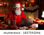 Happy old Santa Claus wearing hat holding gift box using laptop computer sitting at workshop home table late on Merry Christmas eve. Ecommerce website xmas time holiday online shopping e commerce sale