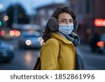 Portrait of happy young woman wearing medical protective mask outdoors and looking at camera. Smiling girl wearing surgical mask for virus protection standing outdoor in winter evening.