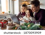Happy young parents with cute excited small kid daughter using laptop computer at home sitting at Christmas table having virtual party on video call, doing ecommerce family online shopping together.