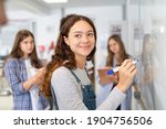Portrait of smiling girl solving math equation on white board. College student thinking and solving arithmetic sum with classmates. Smart young woman writing on white board during class.
