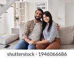 Happy young indian couple real estate buyers hugging sitting on couch at home looking at camera. Smiling husband and wife new homeowners embracing, enjoying own apartment purchase, portrait.