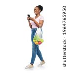 Small photo of sustainability, food shopping and eco friendly concept - happy smiling african american woman with smartphone holding reusable string bag with fruits and vegetables walking over white background