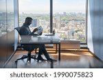 Indian serious investment banker financial analyst sitting at desk with papers working with big data, stock exchange trading operations using laptop near panoramic window in modern corporate office.