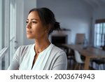 Mature african woman looking outside window with uncertainty. Thoughtful mid adult woman looking away through the window while thinking about her future business after pandemic. Doubtful lady at home.