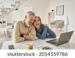Happy mature older family couple laughing, bonding sitting at home table with laptop. Smiling middle aged senior 50s husband and wife having fun satisfied with buying insurance, paying bills online.