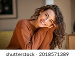 Happy young woman sitting on sofa at home and looking at camera. Portrait of comfortable woman in winter clothes relaxing on armchair. Portrait of beautiful girl smiling and relaxing during autumn.