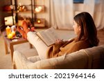 halloween, holidays and leisure concept - young woman reading book and resting her feet on table at cozy home