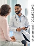 Small photo of Vertical shot of young happy cheerful Indian medical worker therapist in white doctor's robe having appointment consulting older female patient in modern clinic hospital. Medical healthcare concept.