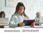 Small photo of Happy smiling teen elementary schoolgirl studying looking at tablet device sitting in classroom with group of schoolchildren using laptop computers. Modern technologies for education concept.