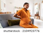 Small photo of Unhappy black expecting woman suffering from lower back pain sitting on sofa with copy space. Tired middle aged african pregnant woman suffer from lower back pain on last month of pregnancy.