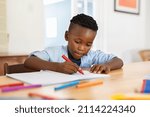 Small photo of Little black boy sitting on table and painting with colored marker on book. Cute african american child drawing in the living room at home. Schoolboy colouring in book with red marker at home.