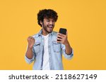 Excited happy young indian man winner feeling joy using smartphone winning lottery game, betting, getting cashback online gift in mobile app message holding cell phone isolated on yellow background.