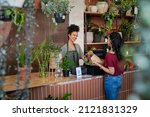 Small photo of Happy black woman entrepreneur standing behind counter in plant store selling fresh flowers to client. Young latin girl buy a fresh bouquet from florist. Smiling african woman botanist selling flowers