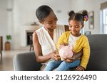 Smiling mature african american mother helping daughter sitting on lap putting money in piggy bank. Cute little black girl saving money by adding a coin in piggy bank with mother at home. 